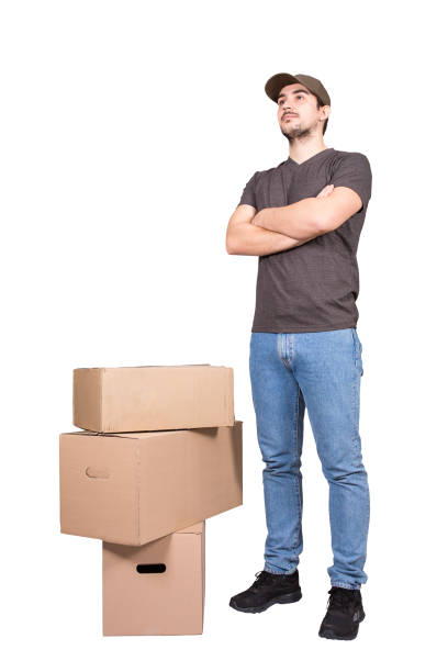 istock Young delivery man wearing cap, full length portrait, stands confident behind the parcel post boxes, isolated on white background. Courier delivering packages. Excellent customer service concept. 1301802463