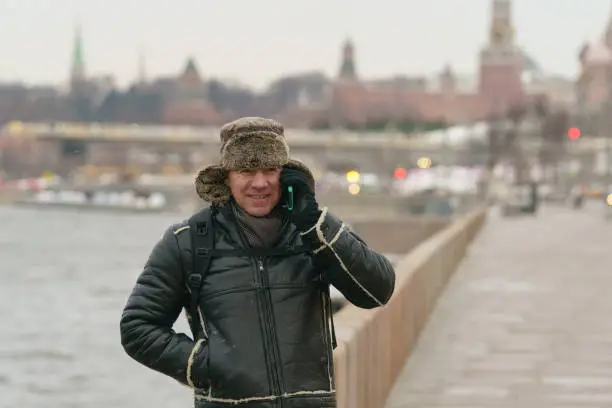 Handsome man using smartphone at the Moscow city street in cold winter day. He wear hot cap made of natural sheepskin. It fastened at the chin to protect from cold. Moscow Kremlin as background.