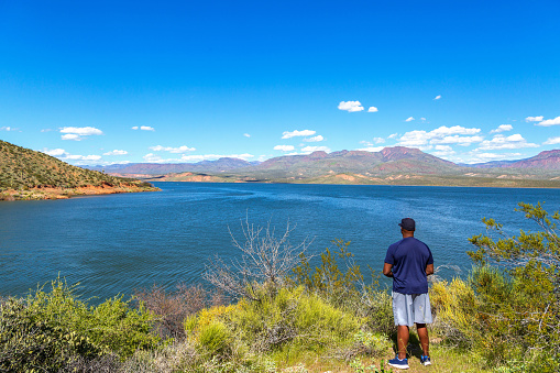 Black man contemplating lake in Arizona's Sonoran Desert in a beautiful sunny day with a blue sky and fluffy white clouds. Many saguaros populate the hills around the lake and come close to the clear blue water of the reservoir.