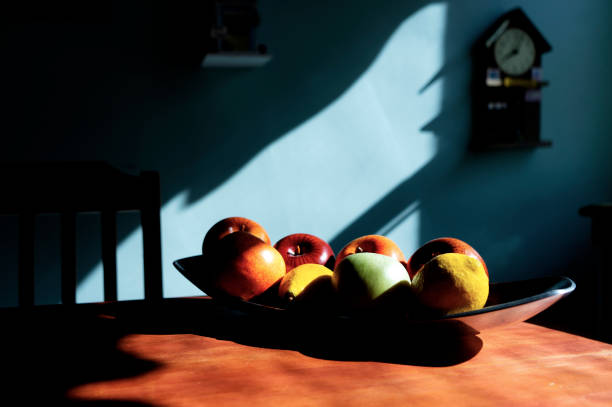 A bowl of wooden fruit - apples and lemon - on a warm wood table, bathed in natural sunlight. A cuckoo clock and slats of a chair complete the scene.