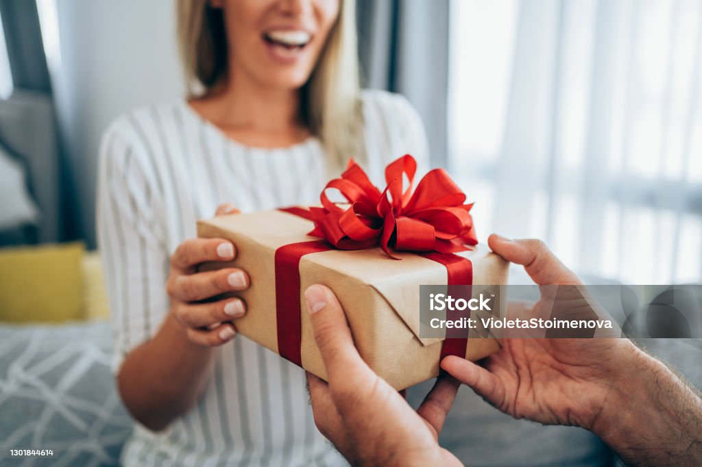 Cheerful young woman receiving a gift from her boyfriend. Cropped shot of a loving husband giving his wife a gift. Boyfriend surprise his beautiful girlfriend with present while she is sitting on the sofa in the living room at home. Focus is on the gift. Gift Stock Photo