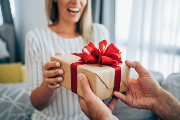 Cheerful young woman receiving a gift from her boyfriend. Cropped shot of a loving husband giving his wife a gift. Boyfriend surprise his beautiful girlfriend with present while she is sitting on the sofa in the living room at home. Focus is on the gift. valentines day holiday stock pictures, royalty-free photos & images