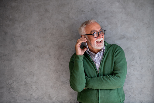 Smiling old man is standing in front of gray wall and talking on his mobile phone with his grandchild. He wears his favorite green sweater and has eyeglasses on.