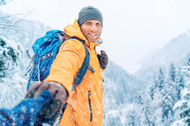 Happy smiling Man dressed bright orange softshell jacket with backpack hand in hand girlfriend point view on the winter mountains route. Active people in nature, help or follow me concept image.