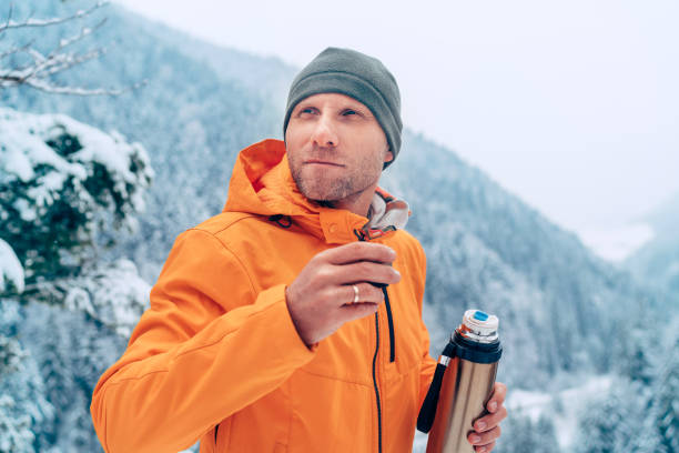 istock Man with a hot drink cup and a thermos flask dressed bright orange softshell jacket drinking while he trekking winter mountains route. Active people in the nature concept image. 1301848244