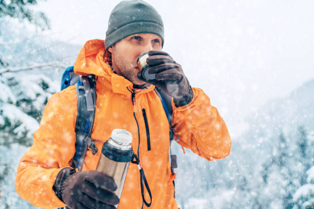 istock Middle-aged Man with a hot drink cup and a thermos flask dressed bright orange softshell jacket drinking while he trekking winter mountains route. Active people in the nature concept image. 1301848248