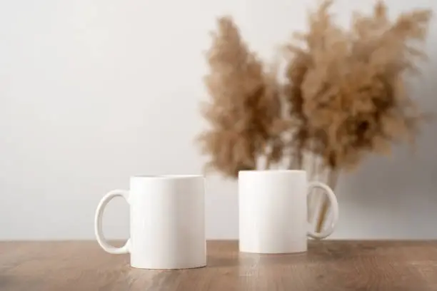 Mockup two white mugs on a wooden table top with pampas decor in a Scandinavian interior, boho style. Template, layout for your design, advertising, logo with copy space. Cups light beige background