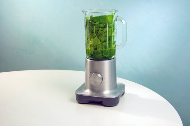 A blender with a glass jug stands on a white table. In the blender lies spinach greens, basil, half-blended into a puree. Vegan food, benefits and vitamins, raw food.