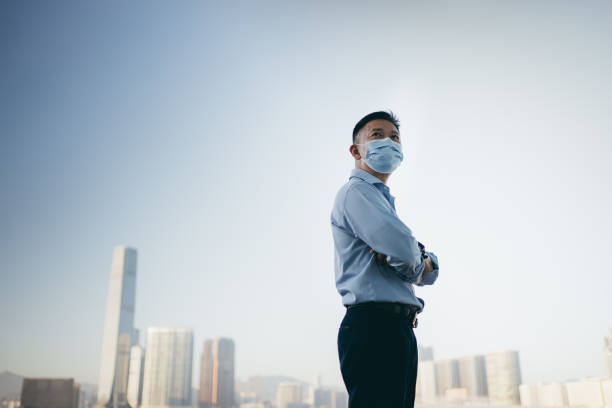 homme d’affaires aîné asiatique utilisant un masque protecteur et regardant vers le ciel traversant des bras par le port de hong kong - senior adult outdoors wellbeing sky photos et images de collection
