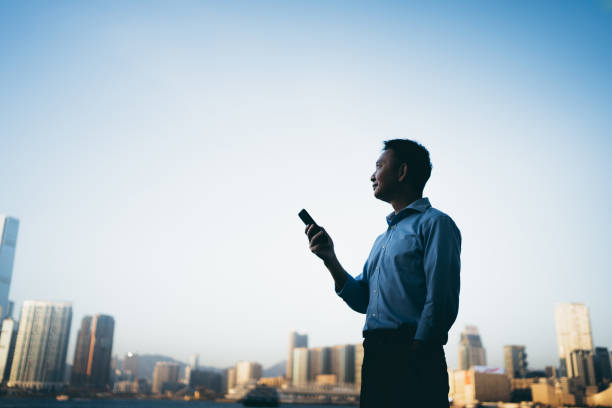 silueta de un hombre de negocios asiático mirando hacia el cielo mientras sostiene su teléfono inteligente junto al puerto de hong kong - hong kong city urban scene building exterior fotografías e imágenes de stock