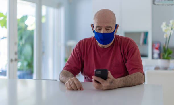 istock Latin senior with a mask sitting at the kitchen counter checking his messages 1301853500