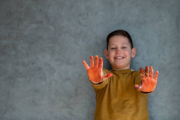Smiling little boy is standing and showing his hands all covered in orange color. Behind him is gray wall and he wears khaki yellow sweater.
