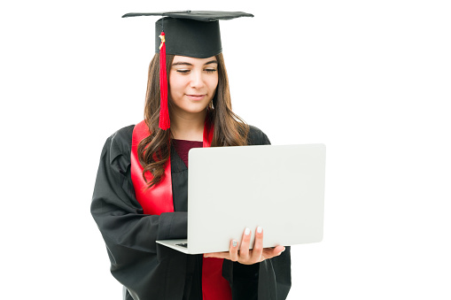 Young female graduate attending via online video call her virtual graduation ceremony while holding the laptop against a white background