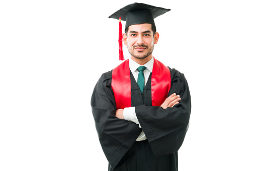 Attractive hispanic man in his 20s wearing a graduation gown and looking at the camera with his arms crossed. Smart guy graduating from university