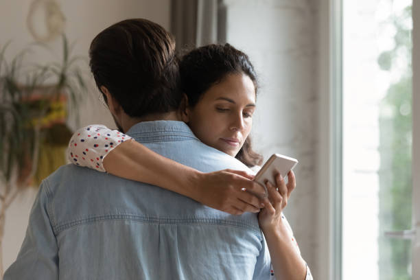 istock Wife hugging husband keeping secret messaging with lover using cell 1301871041