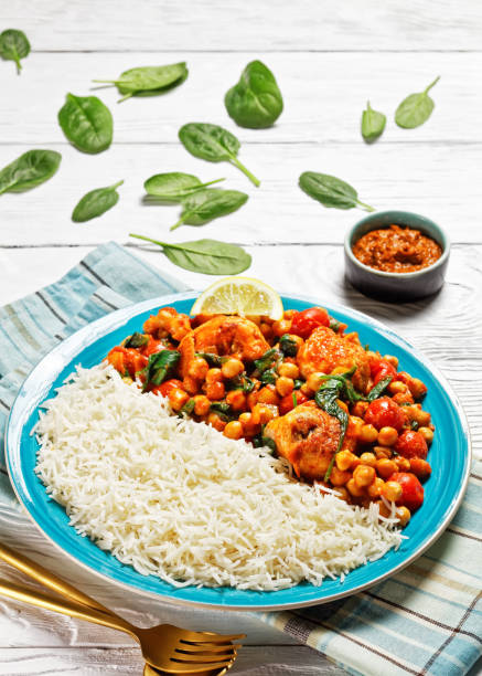 istock Chicken and chickpea curry served with jasmine rice with cherry tomatoes, baby spinach, yellow curry paste served on a blue plate with a lemon wedge on a white wooden background, top view, close-up 1301892522