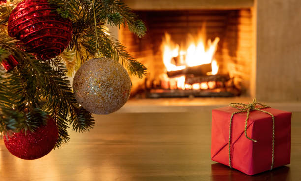 istock Xmas tree branch with balls, red gift box, blur burning fireplace background. 1301895556