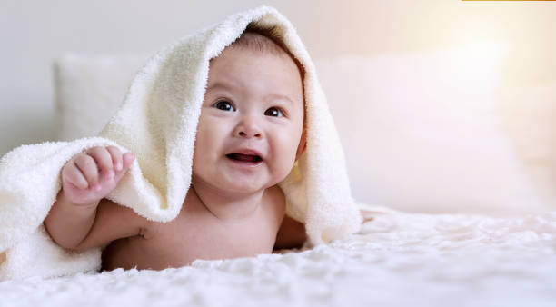 istock Cheerful cute baby looking at camera under white blanket looking at something. Innocence baby crawling on white bed with towel on his head at home. 1301896528