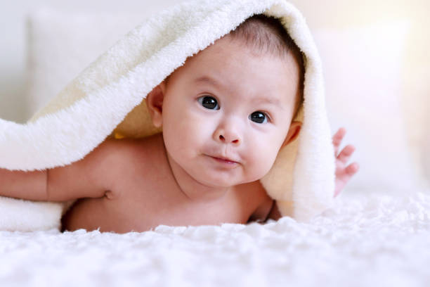 istock Cheerful cute baby looking at camera under white blanket. Innocence baby crawling on white bed with towel on his head at home. 1301896529
