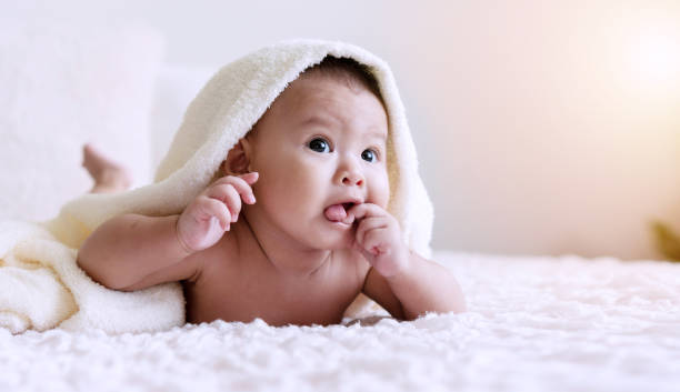 istock Cheerful cute baby looking at camera under white blanket looking at something. Innocence baby crawling on white bed with towel on his head at home. 1301896537