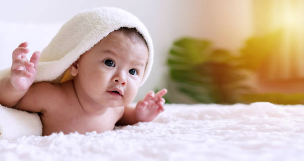 istock Cheerful cute baby looking at camera under white blanket looking at something. Innocence baby crawling on white bed with towel on his head at home. 1301896540
