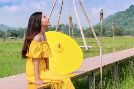 Joyful smile young women wear yellow dresses with yellow umbrella sitting relax on the bridge in the rice field looking at sky. Concept lifestyle relax