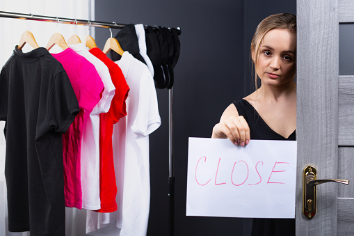 A kind and beautiful blonde, sad, woman working as a consultant in a clothing store indicates that the store is closed on a signboard. Photo indoor shop. High quality photo