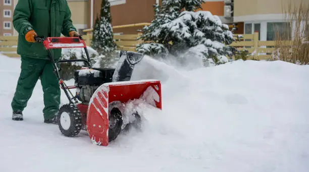 A worker in green overalls removes snow with a red snowblower against the background of a spruce, a snowdrift and a house. High quality photo