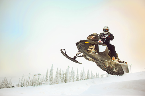 The rider in gear with a helmet making flying jumping taking off on a snowmobile on a background of a winter scenic landscape with mounting hillside and sky.
