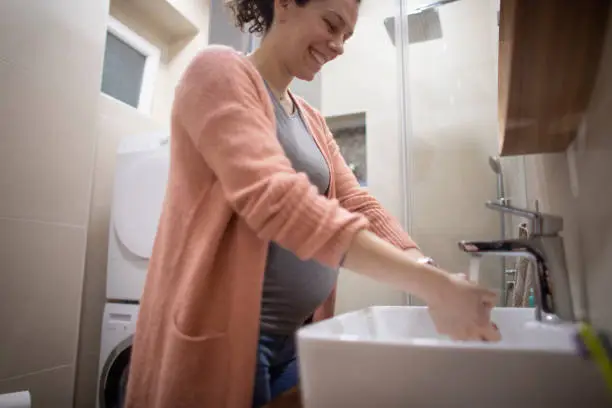 Young pregnant woman washing hands in her bathroom during Covid - 19 pandemic. Focus on hands.