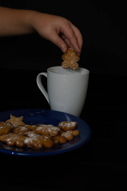 istock Cropped hand picking up cookie from plate on black background and holding white cup- stock photo 1301917367