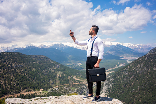 Businessman with mobile phone standing on top of mountain. Young man with white shirt holding mobile phone and business bag.