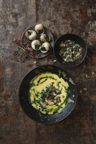 Cream of Pumpkin Soup topped with microgeens, roasted pumpkin seeds and herb green oil. Escargots à la Bourguignonne. Flat lay top-down composition on dark background
