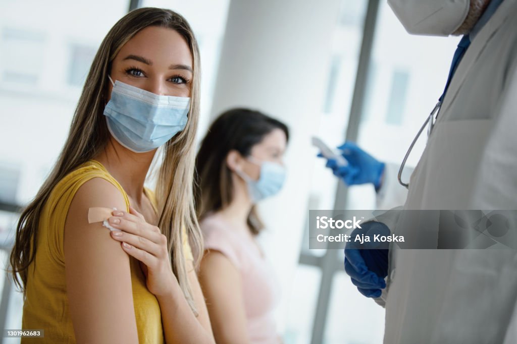 Covid-19 vaccination. Doctor had just vaccinated a young female patient in the hospital. She is pleased with how it all went. She's smiling behind a protective face mask and looking at camera. Medical worker is in protective workwear. Vaccination Stock Photo