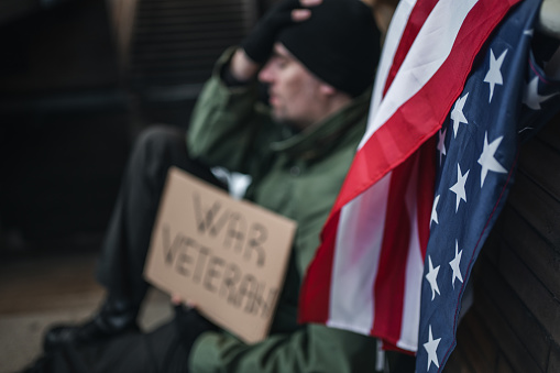 Homeless army veteran sitting on pavement begging on the street holding a placard with text War veteran