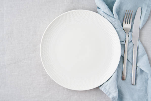 Clean empty white plate, fork and knife on pastel grey linen tablecloth on table, copy space stock photo