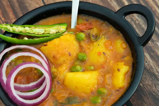 Photo of Top View Of Punjabi Shuddha Shakahari Indian Vegetarian Aloo Matar Ki Masaledar Sabzi Or Bhaji Dipped In Spicy Gravy Curry Served In Traditional Black Kadai Wok And Spoon On Wooden Texture Background