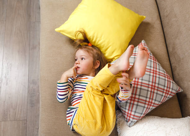 a little girl in yellow trousers and a bright t-shirt is lying on her back on the sofa with her legs up, biting her nails. bad habits in children. - nail biting imagens e fotografias de stock