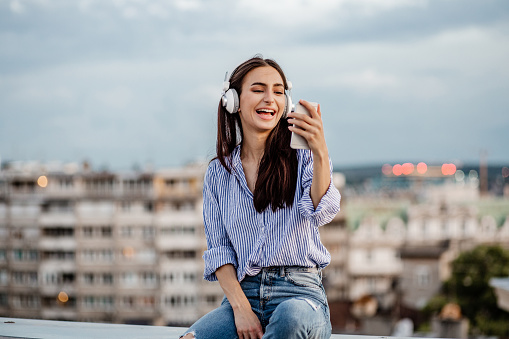 A young woman with headphones is on the rooftop and enjoying herself and listening to music while holding a mobile phone