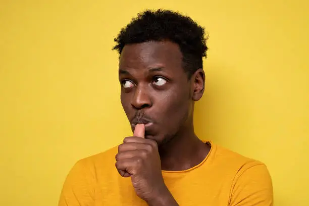 African man with finger in mouth sucking thumb or biting fingernail in anxiety and stress, isolated on yellow background