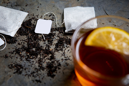 Black tea in transparent cup with slice of lemon is out of focus. On tabletop made of gray stone are white tea bags with white tags and dried leaves of black tea.