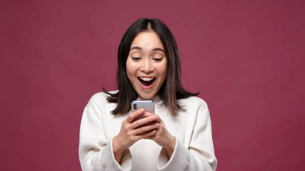 молодая азиатская женщина удивлена телефоном против марсала копировальной области - smart phone asian ethnicity mobile phone women стоковые фото и изображения