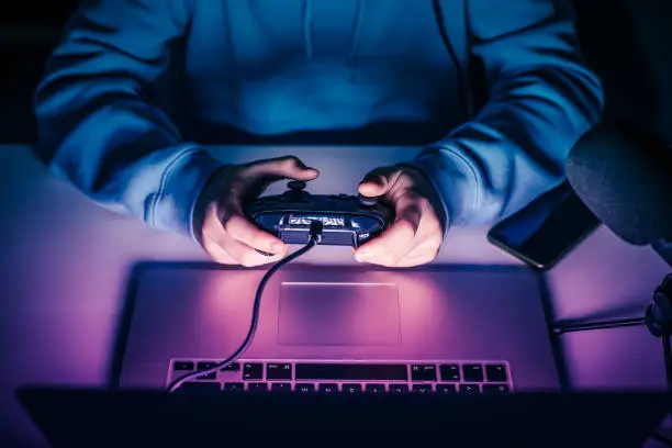 Close up view of young hands using a game pad with laptop to play games online. Details of videogames streamer playing at home console. Vlogger having fun using technology to connect with audiences