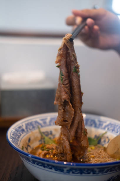 istock Lifting a slice of a premium medium raw Wagyu beef by using chopsticks from Thai noodle bowl. Selective focus 1302029376
