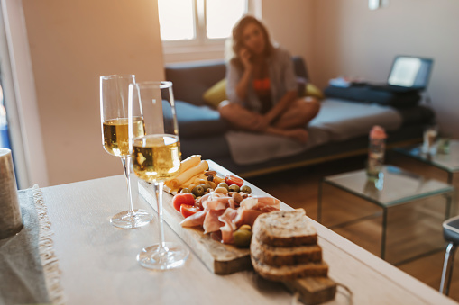 Snacks - olives, meat, cheese, bread and cherry tomatoes on wooden board,  with two glasses of white wine served for a romantic dinner at home, in background young woman sitting on sofa and talking on mobile, calling boyfriend to join her