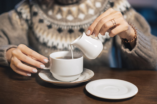 A woman in a winter sweater pours lactose-free milk or cream into a hot drink - coffee or tea. Concept of a cozy cafe and the content of animal fats in the diet