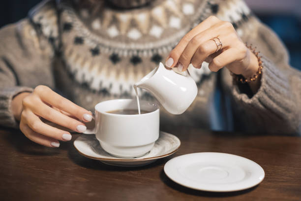a woman in a winter sweater pours lactose-free milk or cream into a hot drink - coffee or tea. concept of a cozy cafe and the content of animal fats in the diet - espresso women cup drink imagens e fotografias de stock