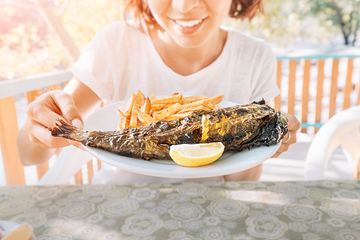 Smiling woman eating fresh river rainbow trout baked according to an old Turkish recipe in a grape or sycamore leaf in a restaurant on a plate with French fries and lemon