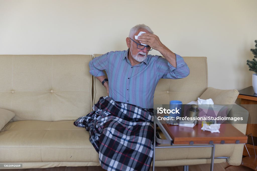 Sick Older Man In The Bed With Seasonal Allergy Is Sweating And Wiping His Forehead With Paper