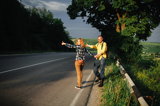 Young happy stylish couple of lovers catch a car in the mountains. Hitchhiking concept.